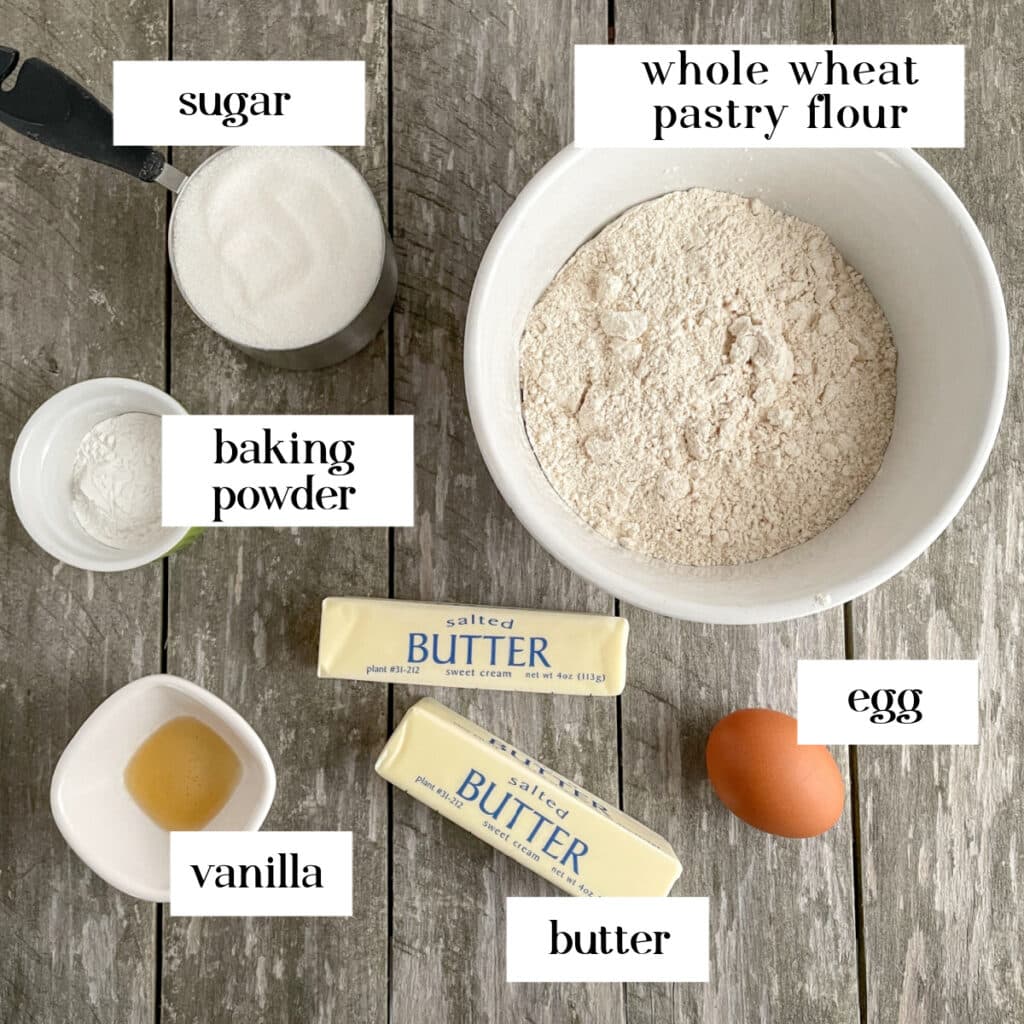 Ingredients for whole wheat Halloween cookies sit on a wood table: sugar, baking powder, vanilla, two sticks of butter, an egg, and a bowl of flour.