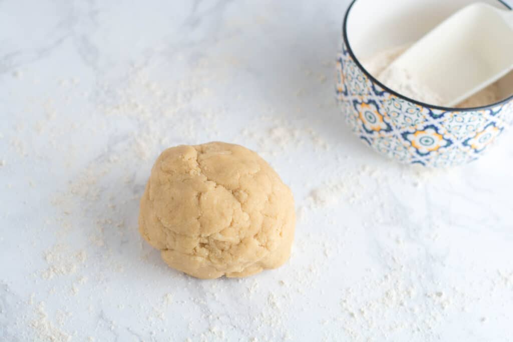 A ball of sugar cookie dough sits on a marble countertop with a bowl of flour next to it.